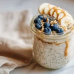 Creamy vegan overnight oats in a glass jar topped with banana, blueberries, and chia seeds.